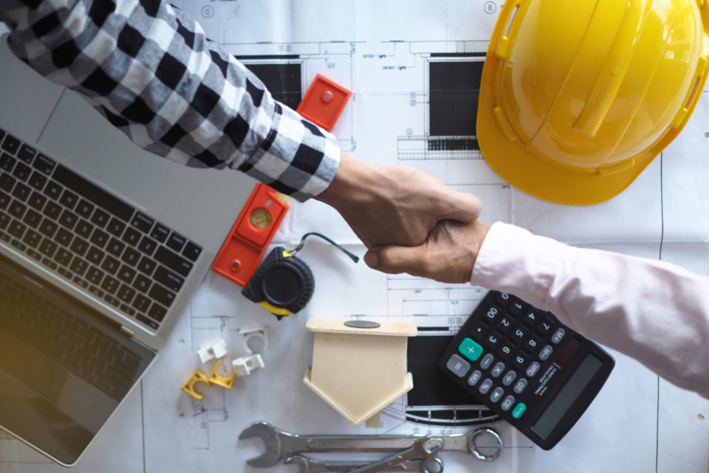 handshake of two persons with yellow color hardhat, calculator, laptop, and construction plan in background